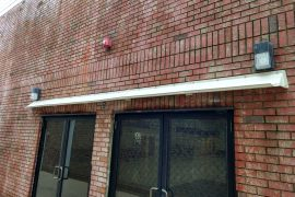 long white DOORBRIM rain diverter installed over 2 bronze sliding glass doors in a brick building.