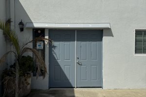 Gray DOORBRIM rain diverter installed above two bluish gray doors and a sidelight to the left in a gray building.