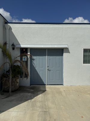 Gray DOORBRIM rain diverter installed above two bluish gray doors and a sidelight to the left in a gray building.