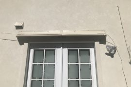 Beige window canopy on second floor above double French doors in beige stucco building.
