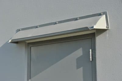 Image of gray DOORBRIM rain diverter mounted over a gray hollow metal door on a gray building.