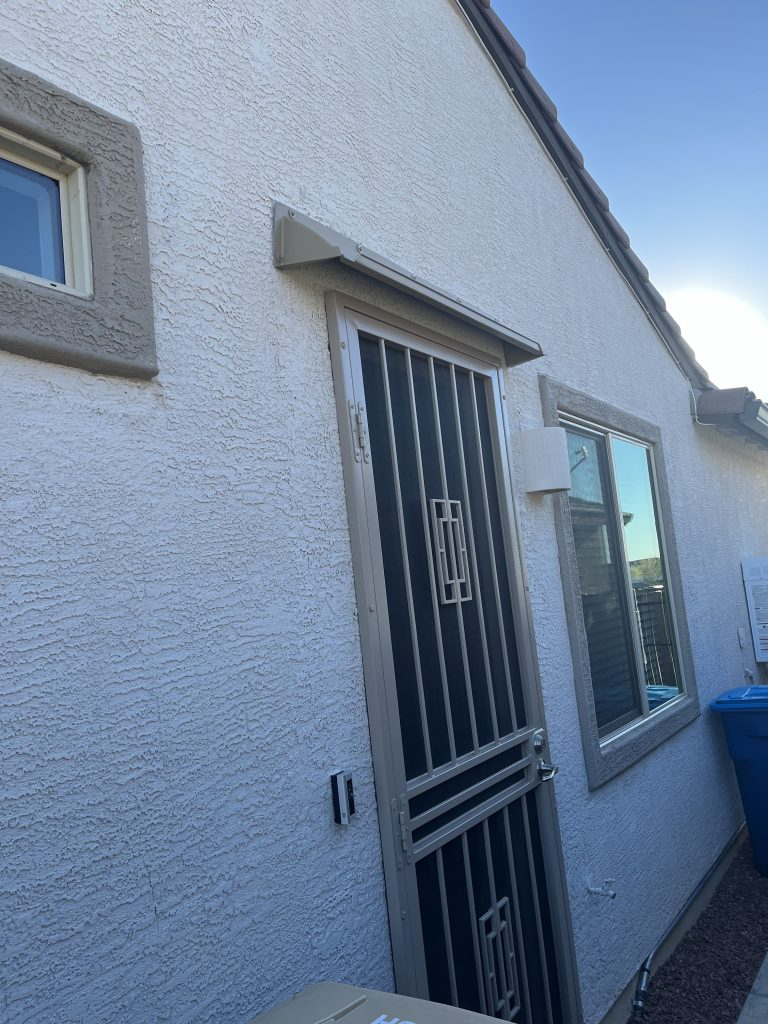 Beige DOORBRIM rain hood mounted over a screen door with vertical bars.