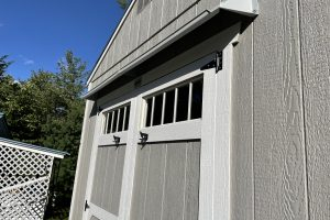 Gray rain guard for shed mounted above double gray and white shed doors with five light panels.