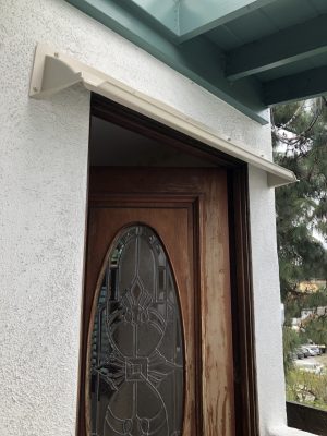 Beige DOORBRIM rain diverter mounted above a dark brown entrance door with an oval glass insert.