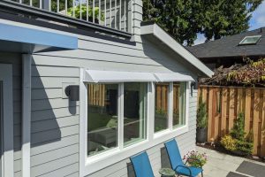 A gray DOORBRIM rain diverter is installed above a large window surrounded by gray clapboard siding for a window leak repair.