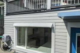 A gray DOORBRIM rain diverter is installed above a large window surrounded by gray clapboard siding for a window leak repair.