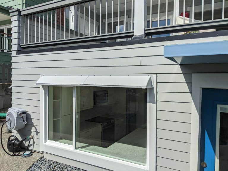 A gray DOORBRIM rain diverter is installed above a large window surrounded by gray clapboard siding for a window leak repair.